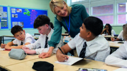 A stock image of a smiling female teacher standing in a bright, modern classroom, engaging a group of students. The students, dressed in matching white shirts and striped black ties, sit at their desks listening attentively.