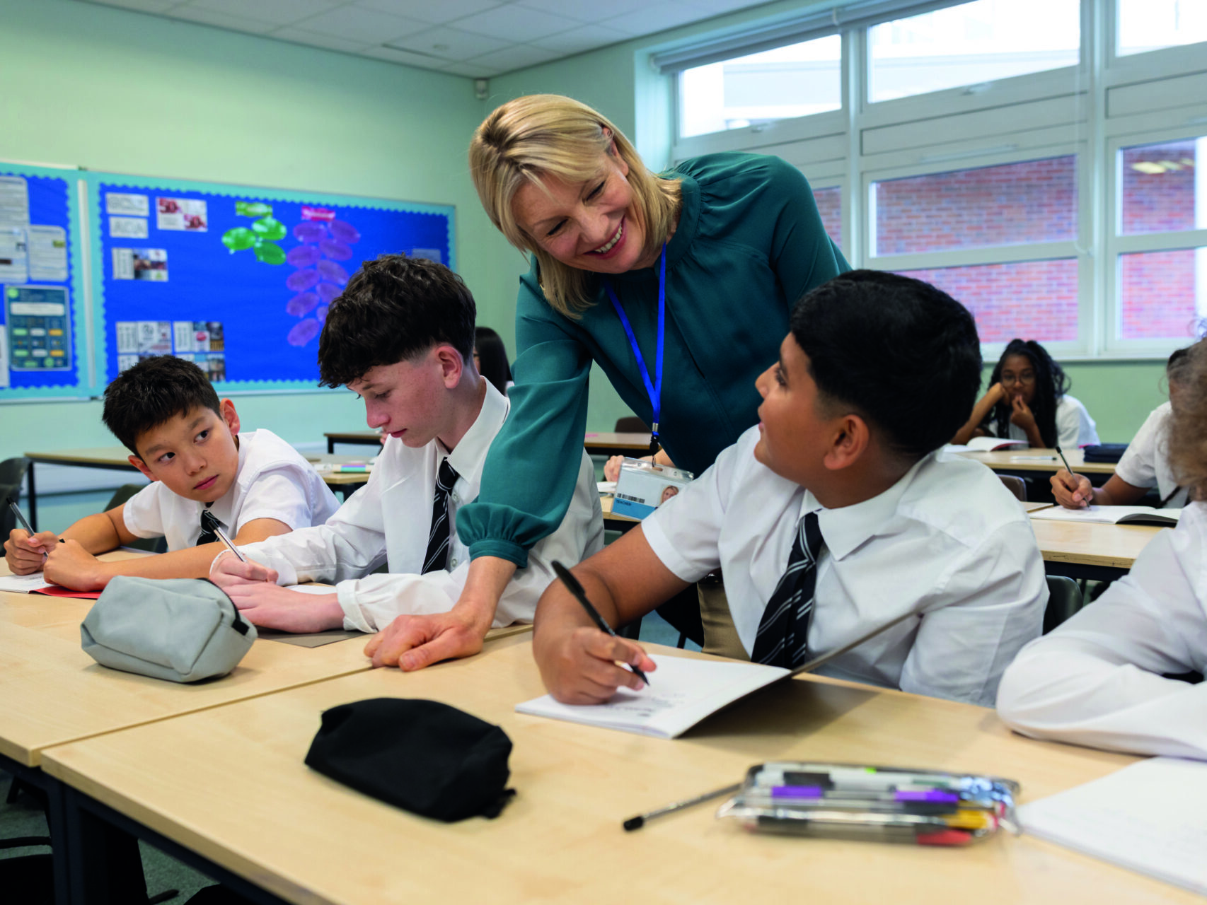 A stock image of a smiling female teacher standing in a bright, modern classroom, engaging a group of students. The students, dressed in matching white shirts and striped black ties, sit at their desks listening attentively.