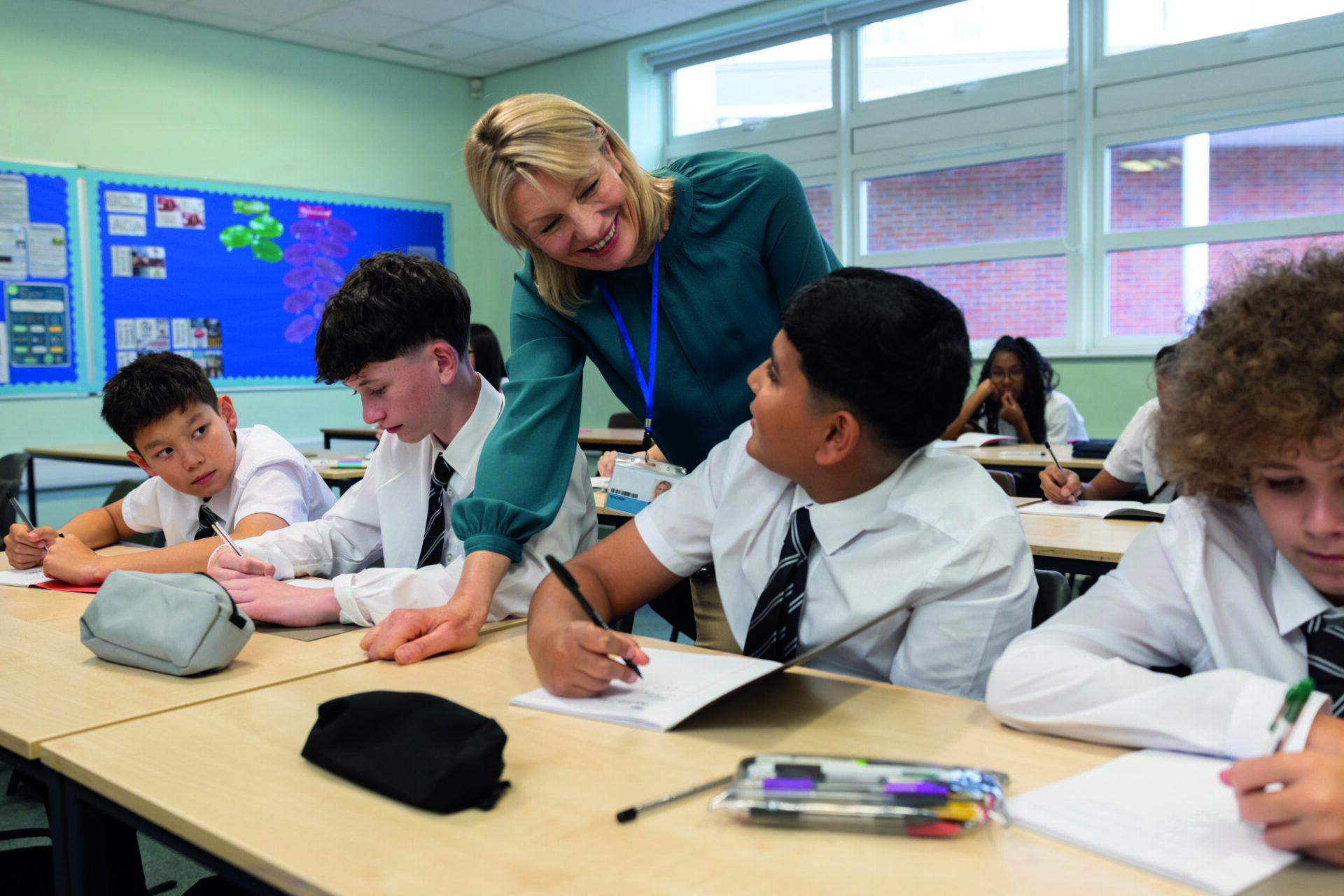 A stock image of a smiling female teacher standing in a bright, modern classroom, engaging a group of students. The students, dressed in matching white shirts and striped black ties, sit at their desks listening attentively.