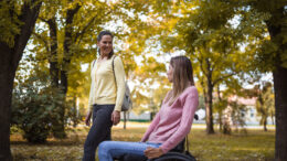 An image of two women spending time together in a park. One of the woman is using a wheelchair. They are smiling and enjoying the autumnal outdoors together.