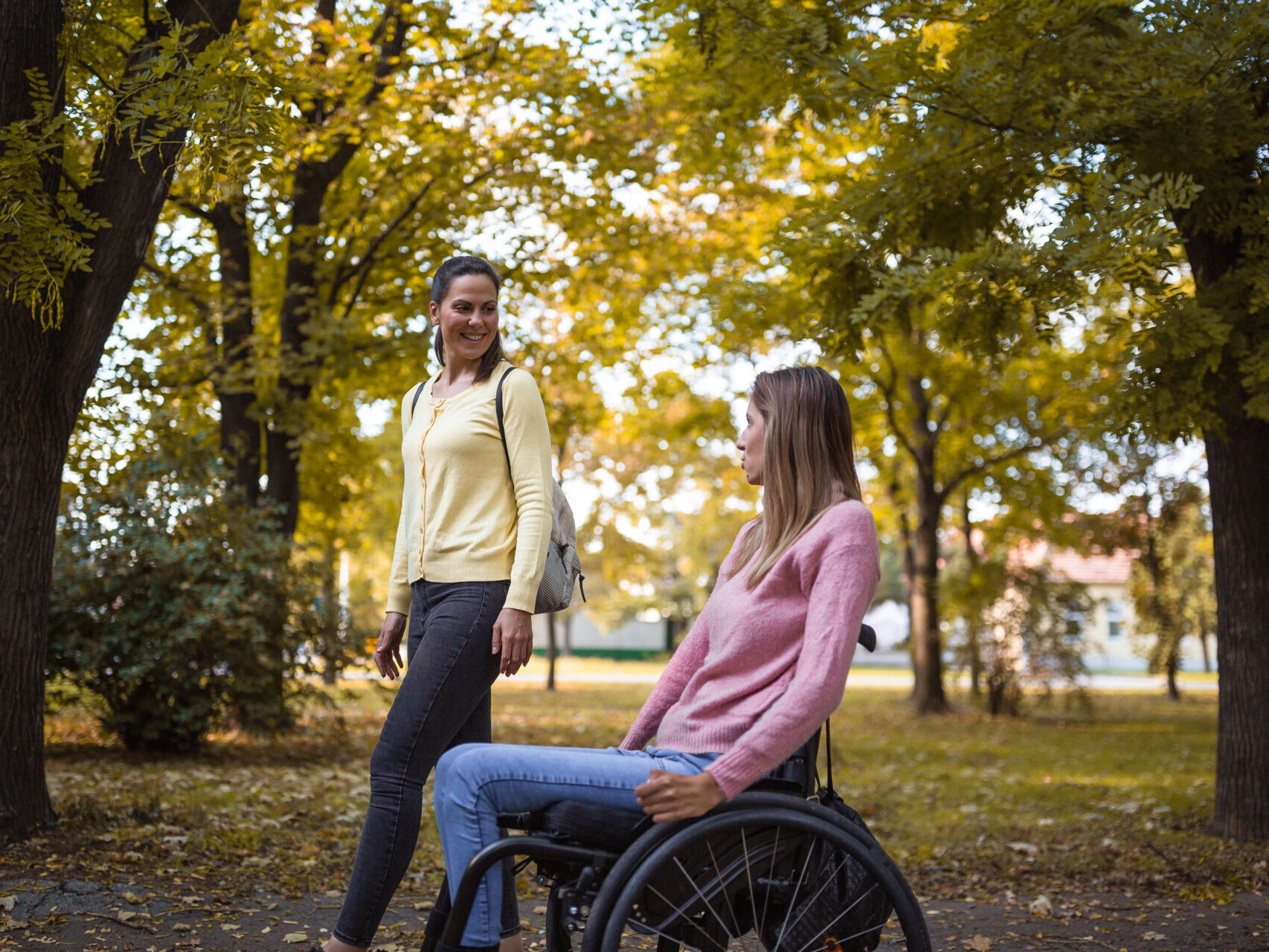 An image of two women spending time together in a park. One of the woman is using a wheelchair. They are smiling and enjoying the autumnal outdoors together.
