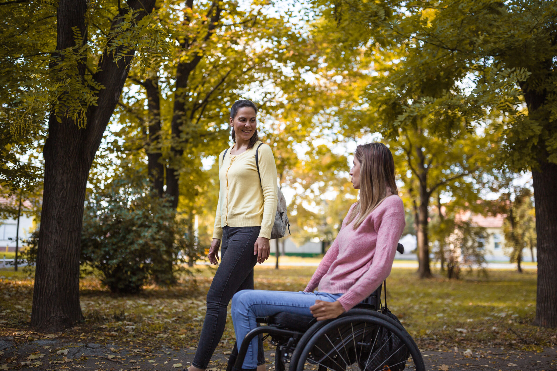 An image of two women spending time together in a park. One of the woman is using a wheelchair. They are smiling and enjoying the autumnal outdoors together.