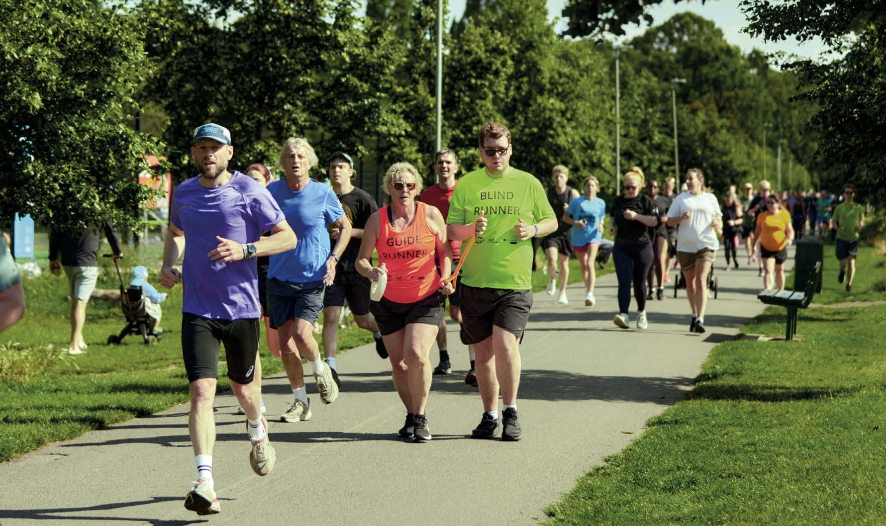 An image of a group of runners at a parkrun. In the centre a visually impaired male runner is taking part, running in tandem with a guide runner to his right. They are wearing coloured t-shirts with 'blind runner' and 'guide runner' labels indicating this.