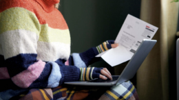 An image of a woman working on her laptop while holding a utility bill. She is wrapped up warm in a stripy turtleneck colourful jumper and fingerless gloves, and a tartan blanket on her legs.