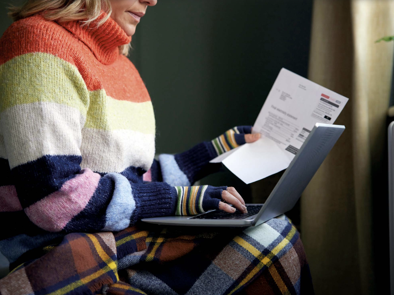 An image of a woman working on her laptop while holding a utility bill. She is wrapped up warm in a stripy turtleneck colourful jumper and fingerless gloves, and a tartan blanket on her legs.